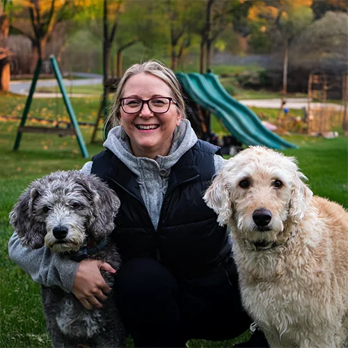 A smiling woman kneeling outdoors with two fluffy dogs, one gray and one cream-colored, in front of a playground.